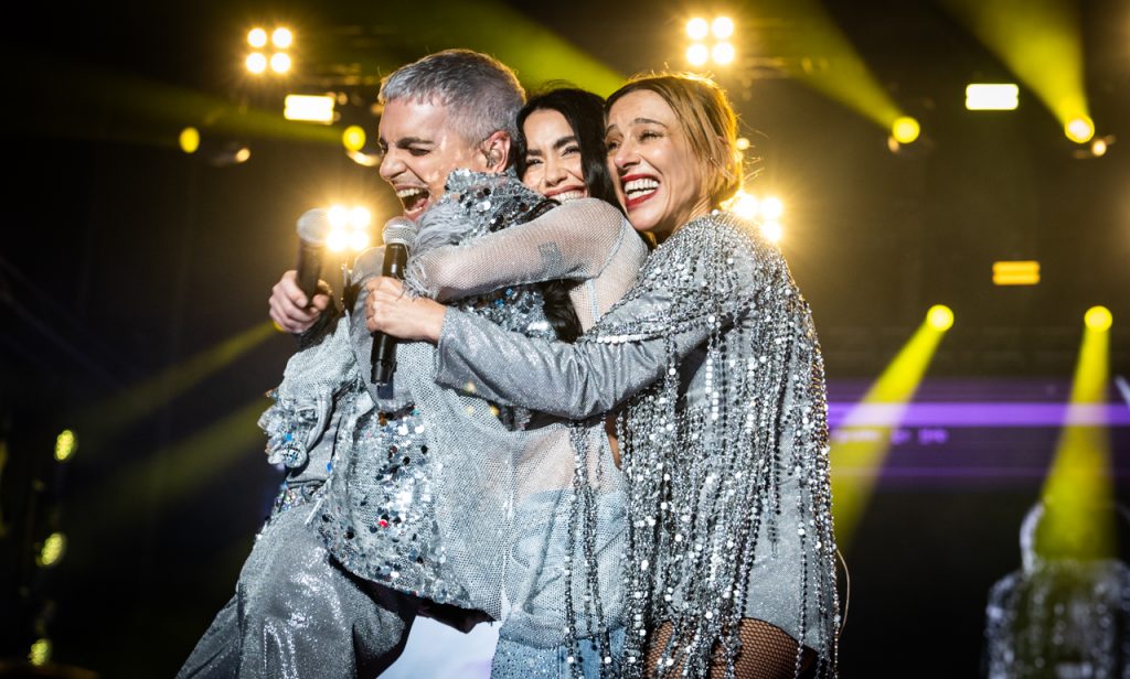 Ale Sergi, Juliana Gattas y Lali abrazados sobre el escenario, los tres con trajes plateados brillantes y las luces amarillas del show detrás.